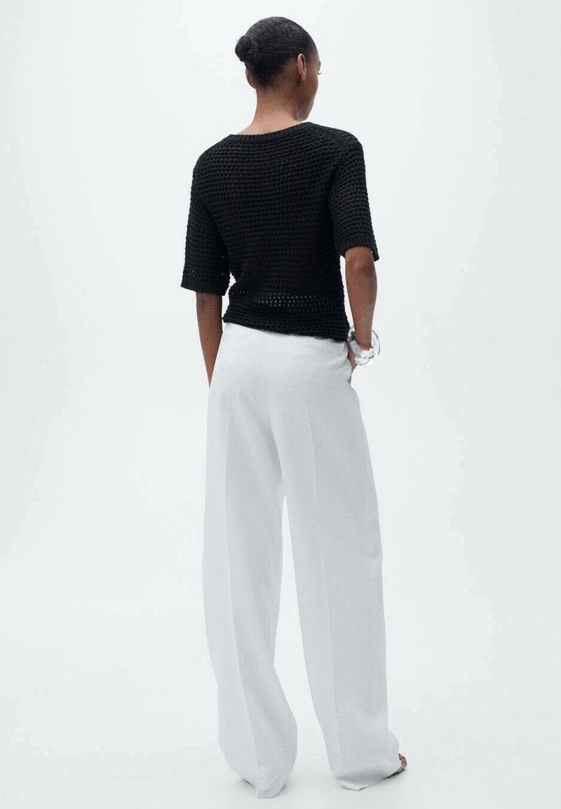 Vrouw met rug naar de camera, gekleed in een zwart is kort mouw gebreid top en witte wijde broek, met handen in de zakken, neutrale achtergrond.