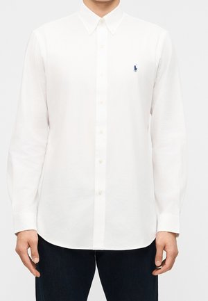 Mand iført en hvid skjorte med lange ærmer og knapper, med en blå broderet polo-spiller logo på venstre bryst.