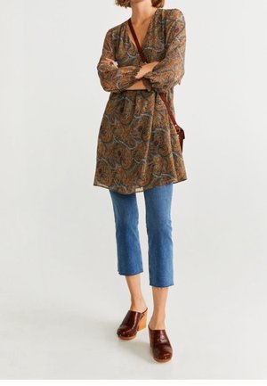 Mujer de pie con los brazos cruzados, vistiendo un vestido marrón de paisley, jeans cortos azules, zuecos marrones y un bolso bandolera.