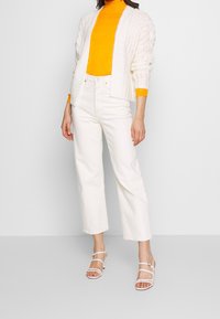 Biały sweter z dzianiny, pomarańczowy golf, białe dżinsy o szerokich nogawkach i białe sandały na paski. Skup się na fakturze dzianiny i kontrastach kolorystycznych.