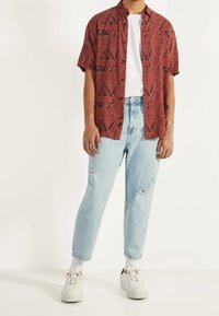 Camisa roja de manga corta con un patrón geométrico negro, combinada con jeans rasgados de color azul claro y zapatillas blancas. Camiseta simple debajo.