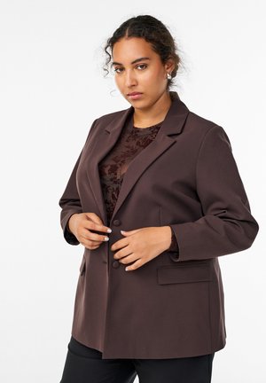 Brun skræddersyet blazer lavet af glat stof, med klapkrave, to knapper og sidelommer. Båret over en gennemsigtig bluse med blomstermønster.