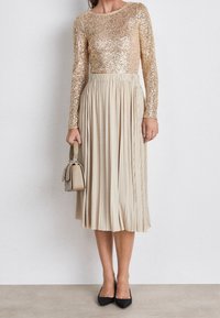 Langlaubige jurk met een top van gouden pailletten en een geplooide beige rok, gecombineerd met een kleine beige tas en zwarte hakken.