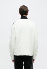 Suéter blanco de gran tamaño y peludo con cuello negro. Tejido texturizado, hombros caídos y dobladillo que termina en la cintura. Detalles en negro a lo largo de las mangas.