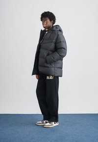 Czarna puchowa kurtka z kapturem, z pikowanym wzorem. Noszona z czarnymi dresami ozdobionymi złotym napisem. Szare i białe sneakersy.