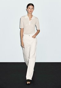 Witte polo met korte mouwen van een textuur gebreid materiaal, gecombineerd met hoge witte broeken. De look wordt afgemaakt met zwarte sandalen met open tenen.