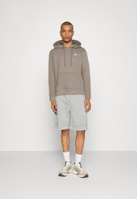 Szürke kapucnis pulcsi elülső zsebbel és zsinórral, világosszürke rövidnadrággal párosítva. A lábbeli bézs és krémszínű árnyalatok keverékéből készült.