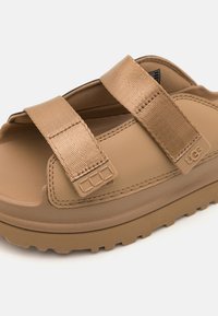 Sandales UGG beiges avec des larges brides texturées, semelle moulée et semelle en caoutchouc épaisse présentant un motif de crampons. Fermeture velcro réglable.