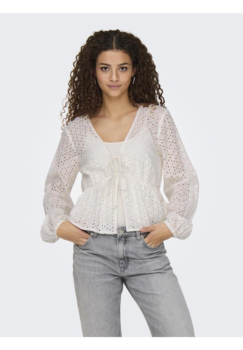 Blusa bianca in pizzo con maniche lunghe, scollo a V e vita con cordoncino. Abbinata a jeans blu chiaro. Presenta un motivo testurizzato.