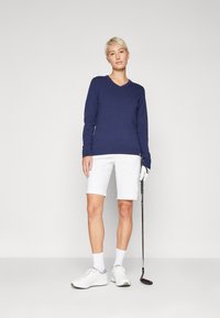 Pull de couleur marine en v, short blanc, baskets blanches, tenant un club de golf. Tissus doux avec des poignets et un ourlet côtelés, design épuré.