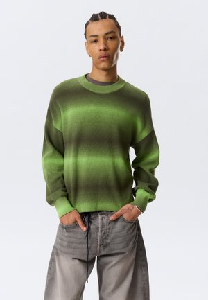 Ung mand med fletninger iført grøn gradienttrøje og løse grå jeans, stående med hænderne i lommerne.