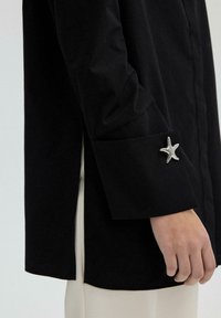 Black shirt with a structured silhouette, featuring a starfish-shaped silver accent on the cuff and a plain texture.