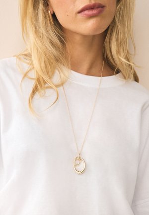 Blonde woman wearing a white shirt and gold necklace with two interlocking oval pendants, against a light background.