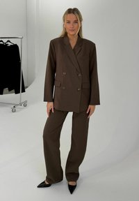 Mujer de pie con traje marrón de doble botonadura y pantalones de pierna ancha, zapatos negros puntiagudos, en estudio blanco con perchero al fondo.