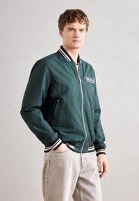 Joven con cabello corto y rizado, vistiendo una chaqueta bomber de color verde oscuro con puños a rayas y jeans gris claro, de pie con una mano en el bolsillo.