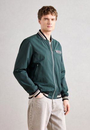 Joven con cabello corto y rizado, vistiendo una chaqueta bomber de color verde oscuro con puños a rayas y jeans gris claro, de pie con una mano en el bolsillo.