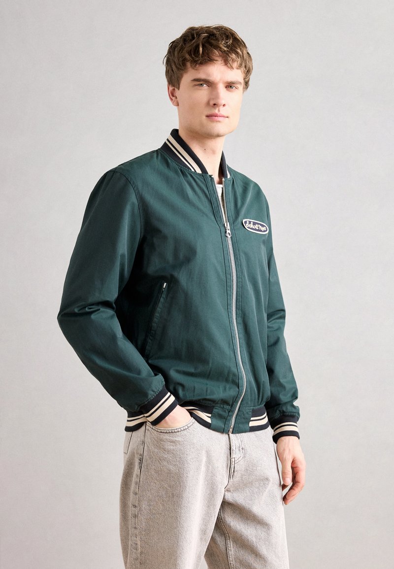 Jeune homme aux cheveux courts et bouclés portant une veste bomber vert foncé avec des poignets rayés et un jean gris clair, se tenant avec une main dans la poche.