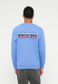 Ανοιχτό μπλε φούτερ με μακριά μανίκια, που διαθέτει γραφική εκτύπωση στο πίσω μέρος, συμπεριλαμβανομένων των "1985" και "TOMMY JEANS" σε έντονες αποχρώσεις.