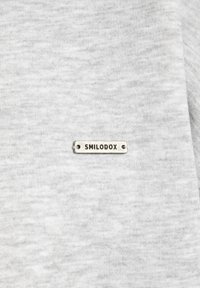 Un tessuto di cotone grigio chiaro presenta un'etichetta rettangolare argento incisa con "SMILODOX", fissata con due piccole viti.