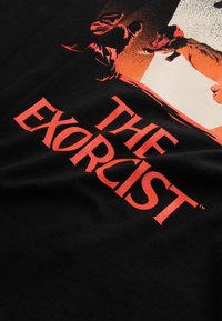 Melnas kokvilnas T-krekls ar grafisku  printu, kurā redzama "Izsistais" ar sarkanu tekstu, kā arī detalizētas figūru un suņa ilustrācijas dažādās toņos.