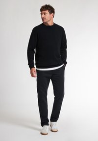 Maglione nero a maglia con scollo rotondo, indossato sopra una maglietta bianca. Abbinato a pantaloni neri e sneaker bianche, in piedi davanti a uno sfondo neutro.