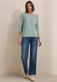 Maglione verde chiaro con scollo a barca e maniche a tre quarti, abbinato a jeans blu a gamba dritta e sneaker bianche.