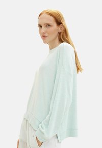 Pulover supradimensionat de culoare albastru deschis, cu margini ribbate și mâneci largi, realizat dintr-un material moale, croi lejer. Modelul stă în profil.