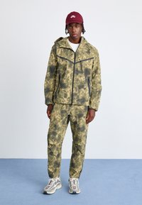 Jachetă și pantaloni cu model camuflat în tonuri de verde și negru, făcuți dintr-un material texturat, cu fermoar frontal și glugă, asortați cu adidași.