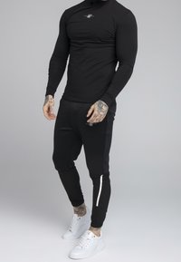 Svart långärmad topp och matchande tapered byxor med vita accenter, tillverkade av elastiskt material. Bärs med vita sneakers. Tatueringar synliga.