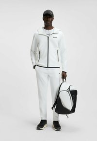 Witte sportieve trainingsoutfit met zwarte accenten, ritszakken en een bijpassende zwart-witte rugzak. Zwarte sneakers maken de look compleet.