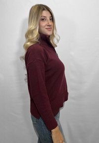 Maglione in maglia borgogna con collo alto e dettaglio con bottoni, caratterizzato da polsini e orlo a coste, indossato con jeans azzurri chiari. Texture liscia.