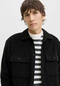 Jeune personne avec des cheveux bruns courts portant des boucles d'oreilles en argent, une veste en velours côtelé noire et une chemise rayée noir et blanc sur un fond uni.