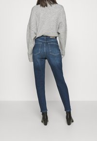 Donna vista di spalle che indossa un maglione grigio, jeans skinny blu scuro a vita alta e stivali neri alla caviglia, in piedi contro uno sfondo neutro.