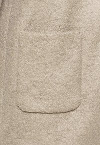 Beige színű filc kabát anyag, puha, texturált felülettel. Egy kerekített szélekkel rendelkező, elülső, egyetlen zsebe található.
