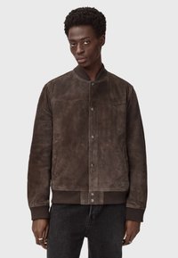 Brązowa zamszowa kurtka bomberka z prążkowanym kołnierzem i mankietami, zapięciem na zatrzask oraz detalami wzdłuż szwu na klatce piersiowej. Nosić ją over czarna dżinsy.