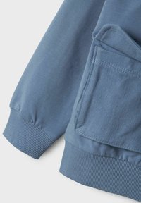 Tela de sudadera azul con una textura suave, que cuenta con puños acanalados y un detalle de bolsillo lateral. El diseño incluye un corte ligeramente holgado.