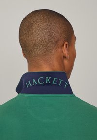 Grønn poloskjorte med en strukturert overflate, med en marineblå krage brodert med "HACKETT" i grønt. Kragen er brettet ned.