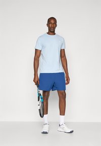 Lyseblå t-shirt med korte ærmer, kombineret med blå sports shorts, hvide sneakers og holder en tennisracket. Simpelt design, uden synlige logoer.