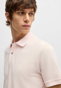 Ανοιχτό ροζ πόλο μπλουζάκι με υφή. Διαθέτει γιακά, κοντά μανίκια και μια διακριτική λογότυπο στο στήθος. Δύο κουμπιά στο άνοιγμα.