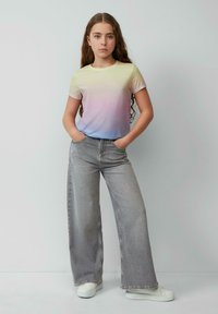 Pastelgradiënt t-shirt dat overgaat van geel naar roze en blauw, gecombineerd met brede lichtgrijze jeans en witte sneakers.