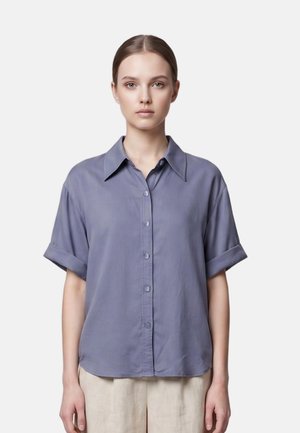 Giovane donna dalla pelle chiara e capelli raccolti indossa una camicia blu larga a maniche corte con bottoni e pantaloni beige, in piedi davanti a uno sfondo semplice.