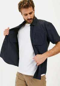 Marineblaues Kurzarmhemd mit Knöpfen auf der Vorderseite und Brusttasche links, getragen über einem weißen T-Shirt, präsentiert mit einer offenen Seite.
