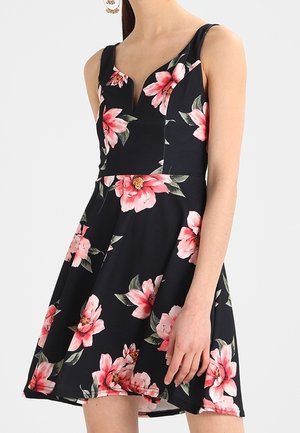 Vrouw draagt een mouwloze zwarte jurk met V-hals en grote roze bloemenprint.