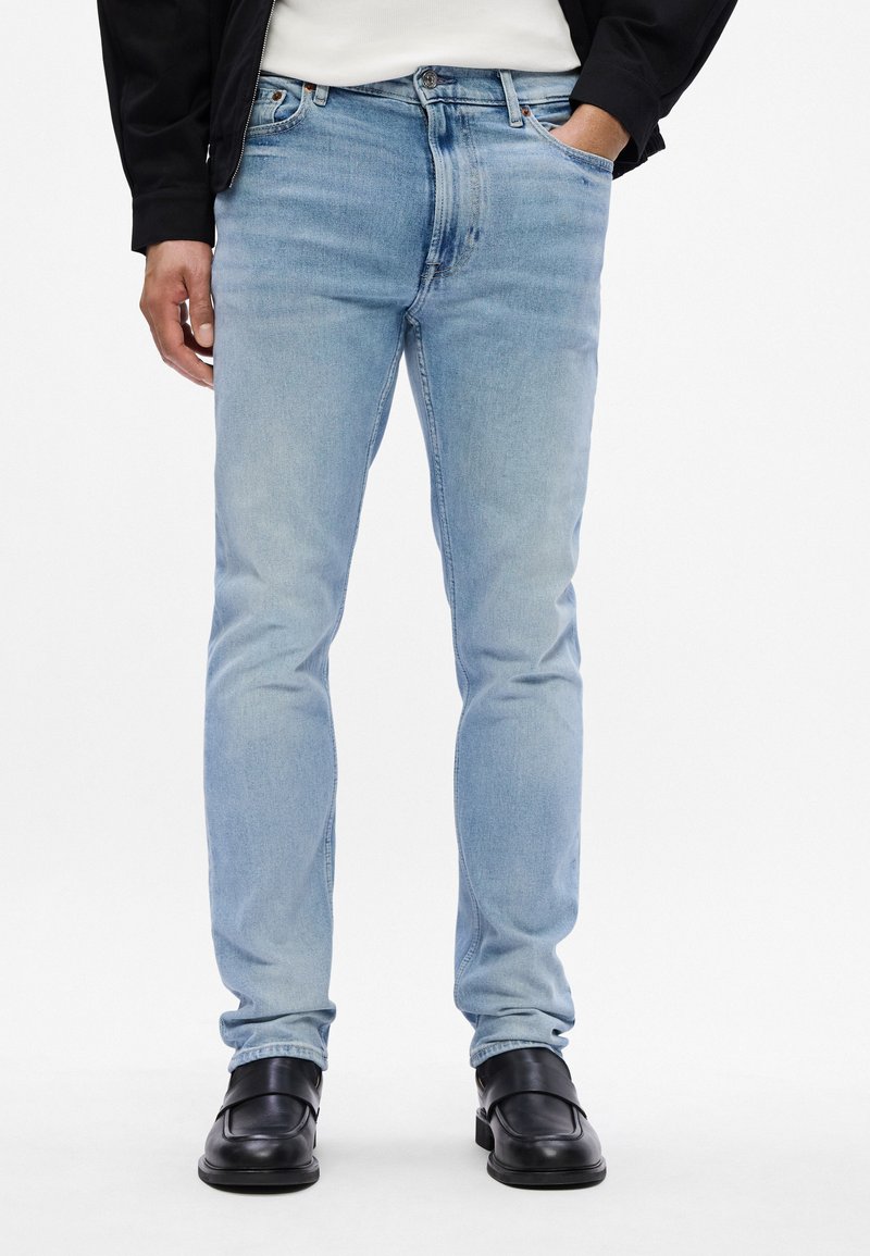 Light blue denim jeans with a fitted design, featuring five pockets and visible stitching. Worn with black slip-on shoes.