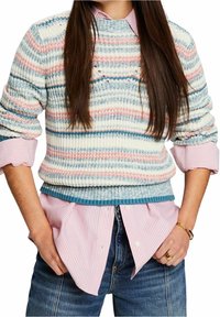 Femme portant un pull en tricot rayé bleu, rose et blanc par-dessus une chemise à rayures rose et blanche, associé à un jean bleu et des bijoux en or.