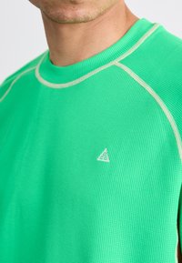 Lyst grøn strikket atletikskjorte med et ribbet stof, der har lys kontrastsyning og et lille trekantet logo på brystet.