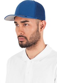 Pălărie albastră cu față solidă și spate din plasă, având o vizieră plată de culoare gri. Pur și simplu peste un tricou polo alb, design minimalist.