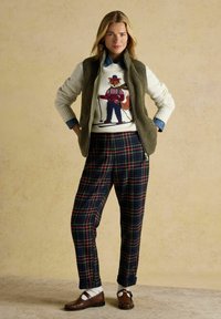 Kārtota apģērba kombinācija, kurā ir krēmkrāsas džemperis ar lapsas dizainu, olīvzaļš veste, svītrainas bikses jūras zila, zaļa un sarkana krāsā, kā arī brūni apavi.