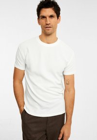 Hvid tekstureret T-shirt med rund hals og korte ærmer, parret med brune bukser, båret af en mandlig model med kort hår og en tatovering på armen.