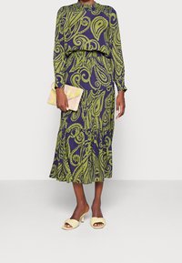 Nainen pukeutuneena pitkähihainen, vihreä ja sininen paisley-kuviollinen mekko, kädessään vaaleanpunainen kukkakuvioinen kirjekuorilaukku ja beigeillä korollisilla sandaaleilla.